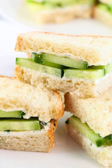3 cucumber and cream cheese sandwiches stacked on a white plate with more sandwiches in the background